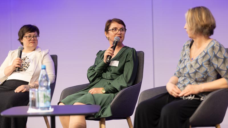 Drei Frauen sitzen auf einer Bühne in Sesseln und sprechen mit Mikrofonen bei einer Podiumsdiskussion.