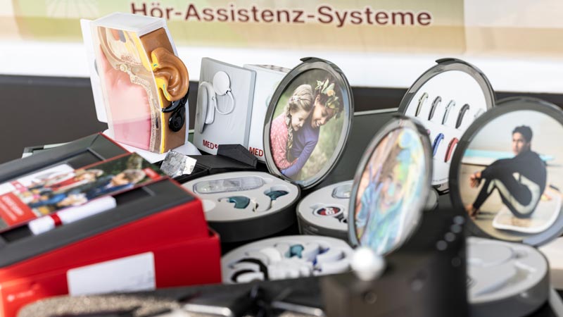 Verschiedene Hörassistenzsysteme und Hörgeräte in Auslagen mit Demo-Ohrmodell und Produktverpackungen.