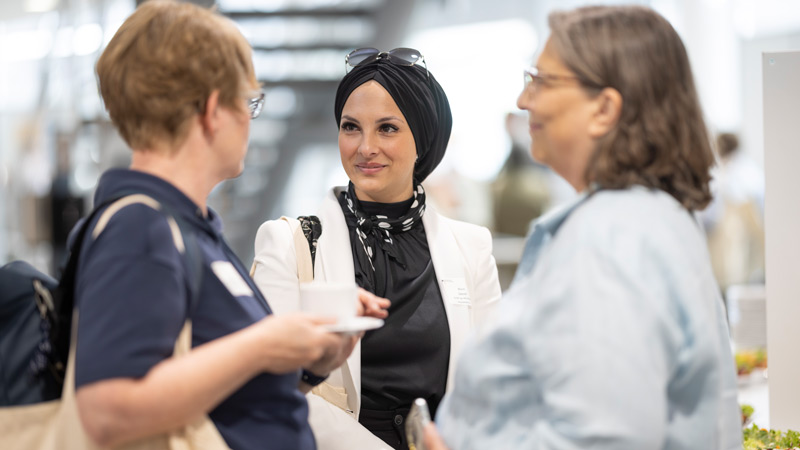 Drei Frauen im Gespräch bei einer Tagung, darunter eine Frau mit schwarzem Kopftuch und weißem Blazer.