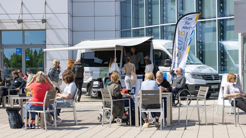 Menschen sitzen draußen an Tischen vor einem Infomobil, darunter auch Rollstuhlnutzende; sonniges Wetter, Infostand mit Fahnen.
