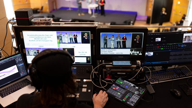 Technikregie mit mehreren Monitoren, die eine Bühne mit Gebärdensprachdolmetschung und Livestream-Anzeige zeigen.