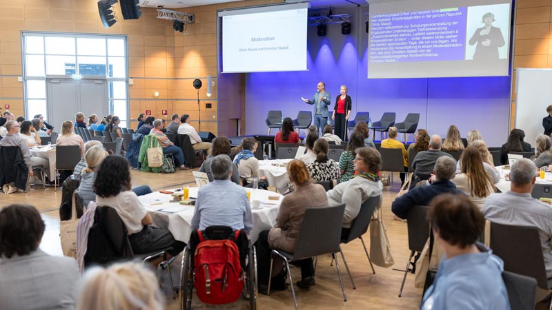 Große Veranstaltung in einem Saal mit Bühne; zwei Moderator*innen sprechen, Gebärdensprachdolmetschung auf Leinwand sichtbar.
