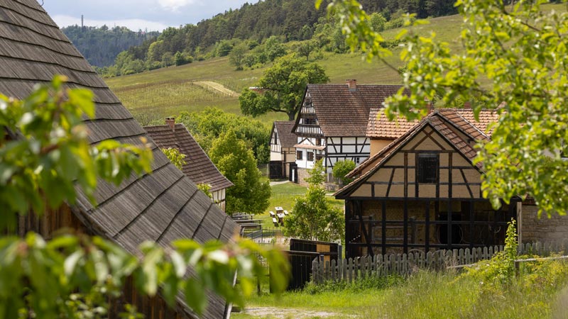 Blick über mehrere traditionelle Fachwerkhäuser in einem Museumsdorf in hügeliger Landschaft.