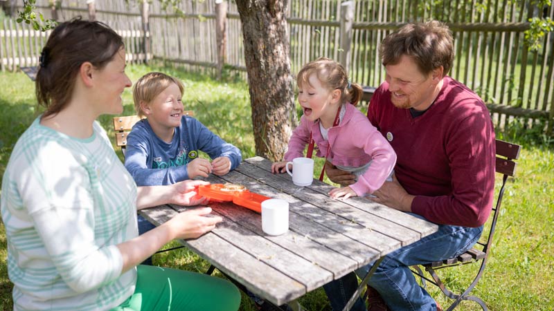 Familie sitzt lachend am Holztisch im Garten und genießt gemeinsam eine Mahlzeit im Freien.
