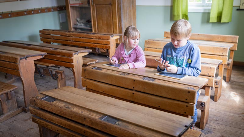 Zwei Kinder sitzen in einer alten Schulbank und schreiben auf kleinen Schiefertafeln.