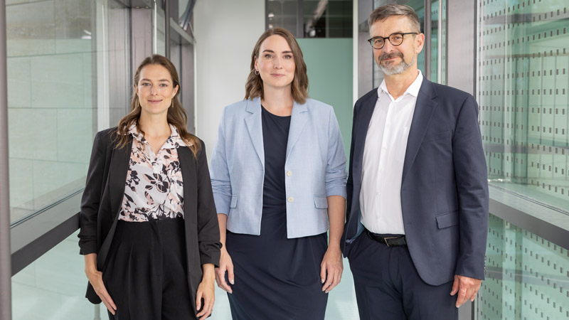 Drei Personen in Businesskleidung stehen nebeneinander in einem modernen Büroflur, alle schauen professionell in die Kamera.