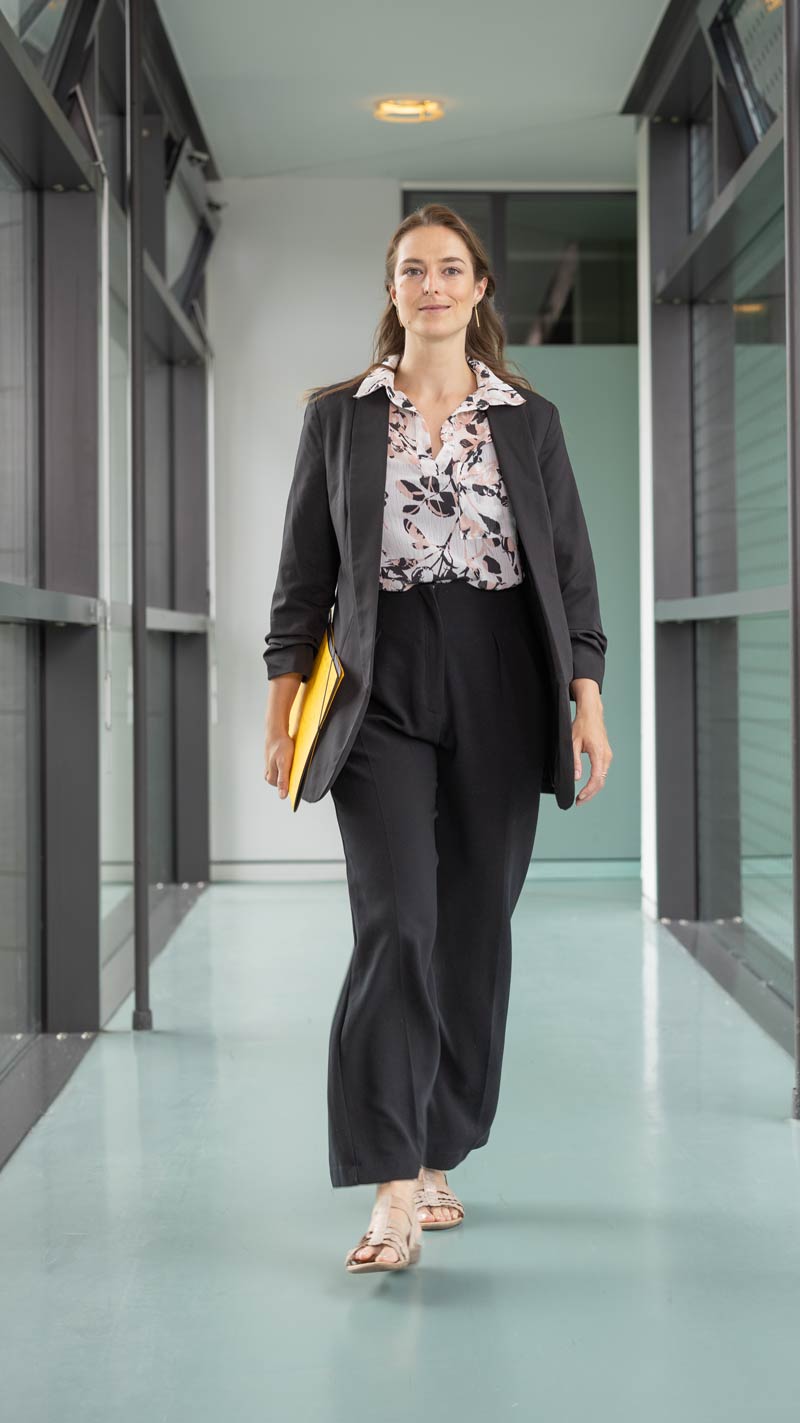 Frau in schwarzem Blazer und gemusterter Bluse läuft selbstbewusst durch einen modernen Büroflur, gelbe Mappe in der Hand.