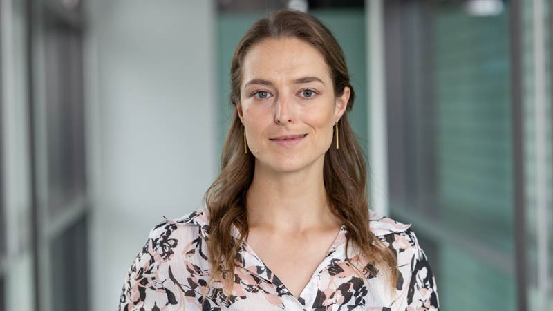 Porträt einer Frau in gemusterter Bluse, frontal vor einer Glaswand mit Lichtreflexionen, direkter Blick in die Kamera.