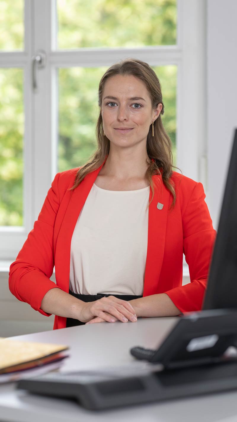 Junge Frau im roten Blazer sitzt am Schreibtisch vor einem Fenster, Blick in die Kamera, selbstbewusst und freundlich.