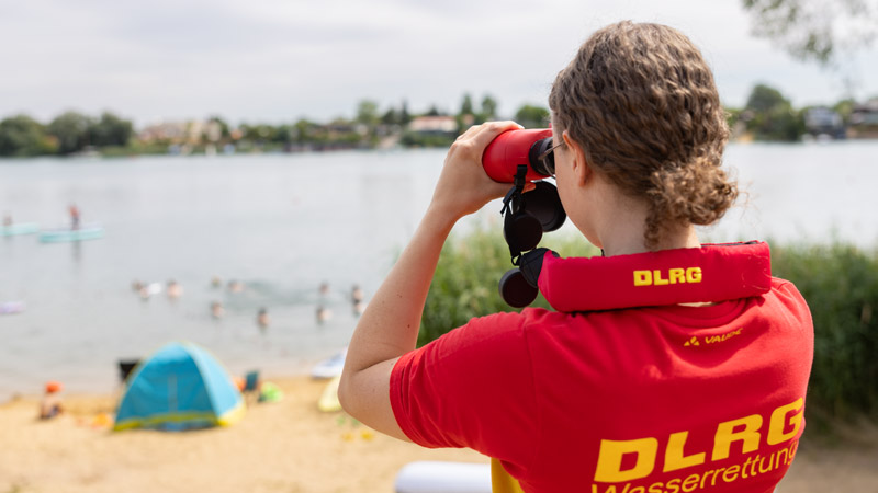 Rettungsschwimmerin mit DLRG-Shirt beobachtet Badegäste am Seeufer durch ein Fernglas.