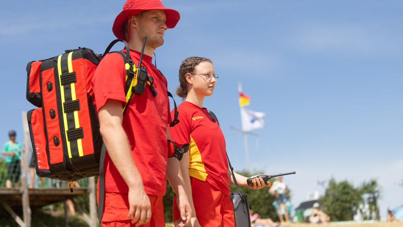 Zwei DLRG-Rettungskräfte in Rot-Gelb-Uniformen stehen am Strand mit Funkgeräten und Notfallrucksack.