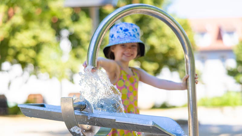 Mädchen mit blau-weißem Hut spielt lachend an einem Wasserspielplatz, Wasser spritzt aus einer Metallkonstruktion.