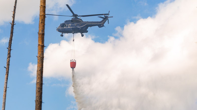 Polizei-Helikopter wirft Wasser aus einem Löschbehälter, Rauch oder Dampf im Hintergrund.