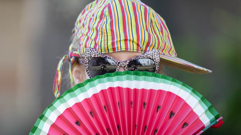 Person mit bunter gestreifter Kappe und Leoparden-Sonnenbrille hält einen Fächer im Wassermelonen-Design vor dem Gesicht.