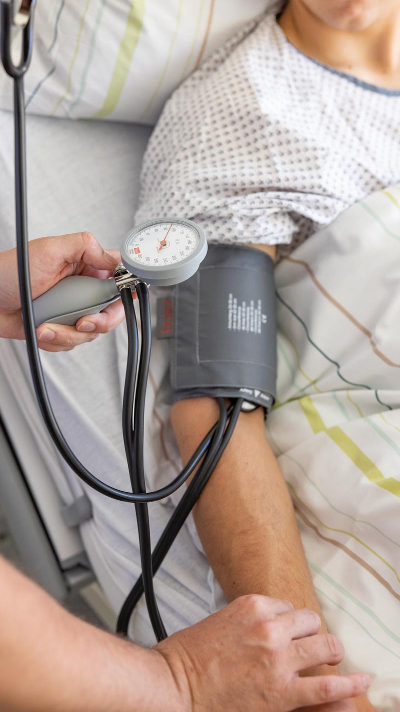 Nahaufnahme einer Blutdruckmessung mit einer Manschette am Arm eines liegenden Patienten.