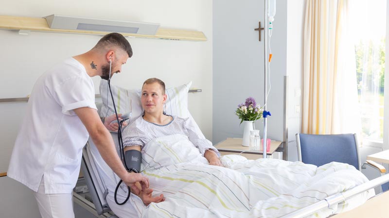 Pfleger misst Blutdruck bei einem Patienten im Krankenhausbett, beide lächeln sich an.