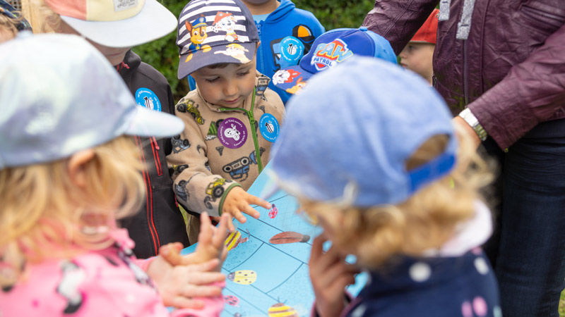 Gruppe von Kindern mit Mützen spielt an einem Tischspiel mit Insektenmotiven.