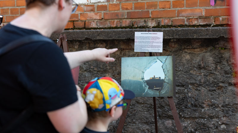 Erwachsener und Kind mit bunter Mütze betrachten ein Foto mit erklärendem Text bei einer Outdoor-Ausstellung.