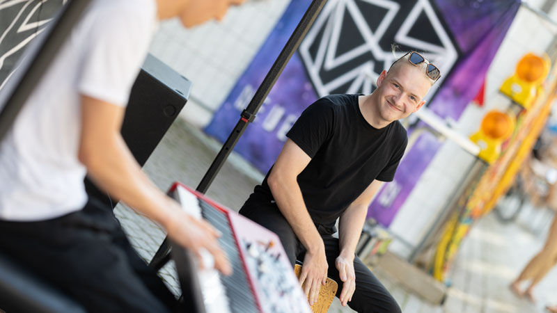Straßenmusiker spielt Cajón und lächelt, während ein anderer Keyboard spielt, Festivalbanner im Hintergrund.