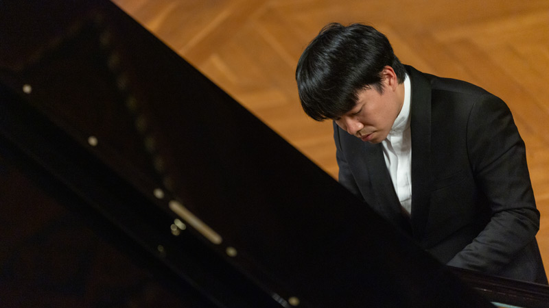 Konzertpianist im schwarzen Anzug spielt konzentriert auf einem Flügel auf Parkettboden.