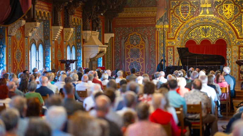Publikum in prunkvollem Saal mit Wandmalereien und Flügel, gespannt auf ein klassisches Konzert.