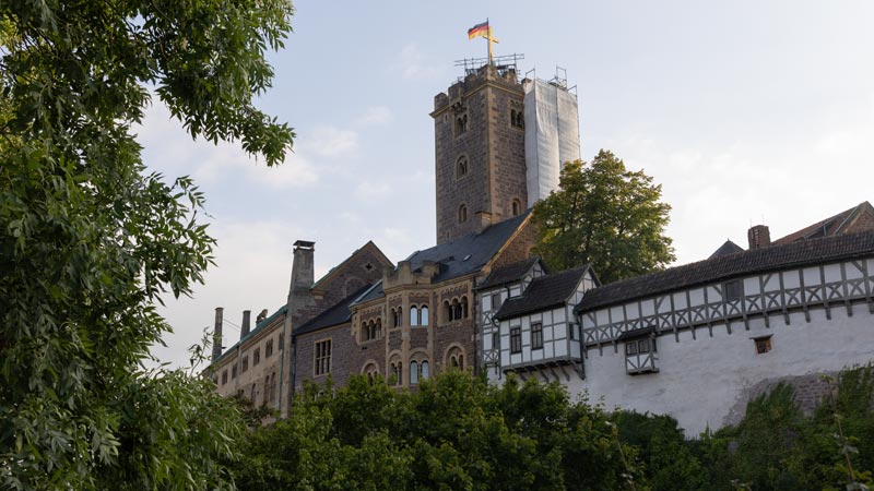 Die Wartburg bei Eisenach mit deutscher Flagge auf dem Turm, umgeben von Bäumen und Fachwerkgebäuden.