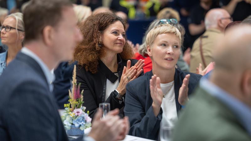 Menschen sitzen an Tischen und applaudieren während einer Veranstaltung.