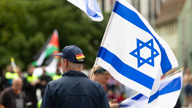 Mann mit Mütze in Deutschlandfarben hält eine israelische Fahne, im Hintergrund unscharf palästinensische Flaggen.