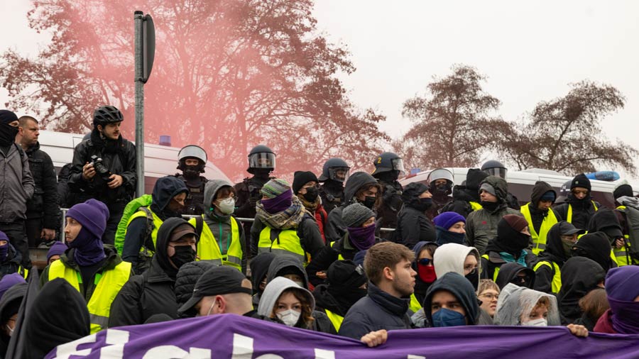 Große Gruppe vermummter Demonstrierender mit gelben Westen vor Polizeikräften und rotem Rauch im Hintergrund