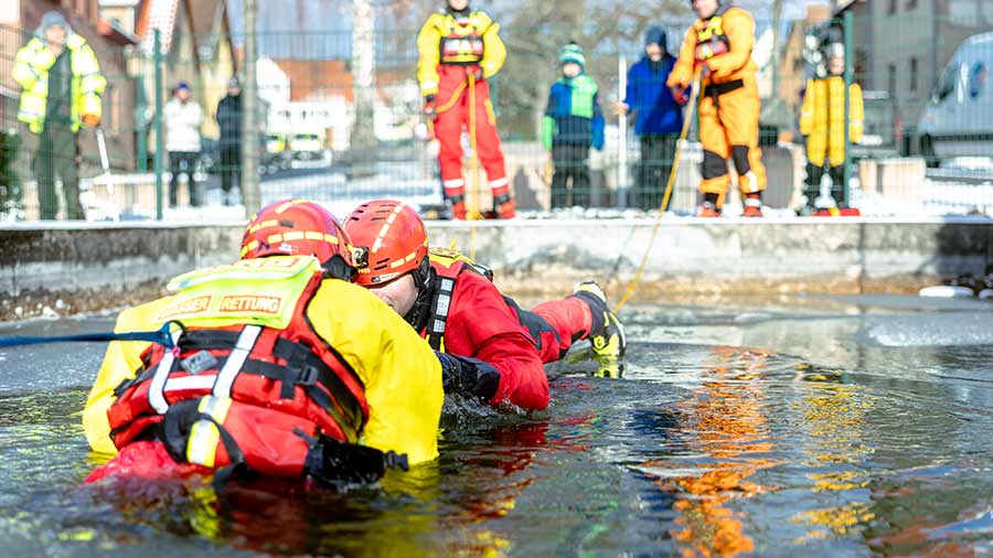 Zwei Einsatzkräfte in Eisrettungsanzügen ziehen eine Person aus dem eiskalten Wasser.