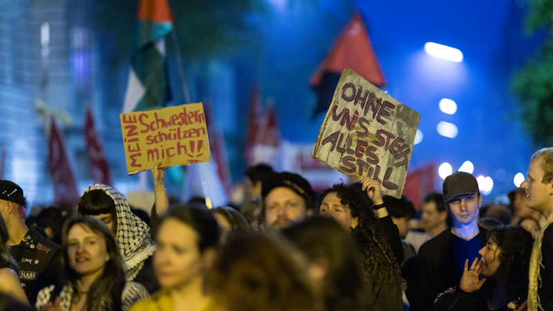Nachtaufnahme der Mai-Demo in Berlin: Teilnehmende halten handgeschriebene Schilder, blaues Licht der Straßenbeleuchtung