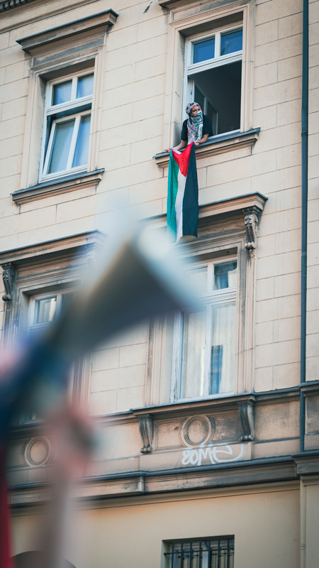 Person hält palästinensische Flagge aus einem Fenster in Berlin-Kreuzberg während der Mai-Demo, im Vordergrund verschwommenes Megafon.