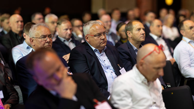 Konzentriertes Publikum bei einer Fachtagung: Mehrere Reihen Teilnehmender in Businesskleidung verfolgen aufmerksam die Bühne. Ein Mann mit roter Brille schaut direkt in die Kamera. Konferenzatmosphäre mit dunklem, warmem Hintergrundlicht.