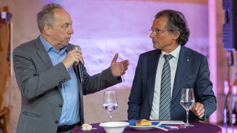 Zwei Männer in Jackett im Gespräch. Der linke trägt eine schwarze Brille. Heller, unscharfer Hintergrund. Netzwerken in der Veranstaltungspause.