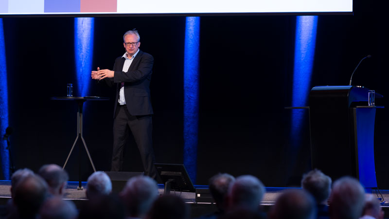Ein Mann im Jacket steht hinter einem Rednerpult und spricht ins Mikrofon. Sein Gesicht ist scharf, das Publikum im Vorder- und Hintergrund unscharf. Blaues Bühnen- und Konferenzlicht.