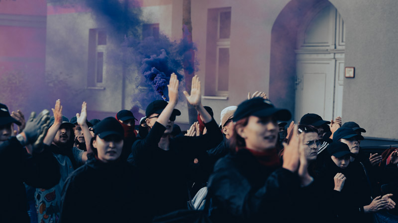 Gruppe von Demonstrierenden mit erhobenen Händen vor einem historischen Gebäude, im Hintergrund lila Rauch einer Bengalfackel, gedämpfte Lichtstimmung.