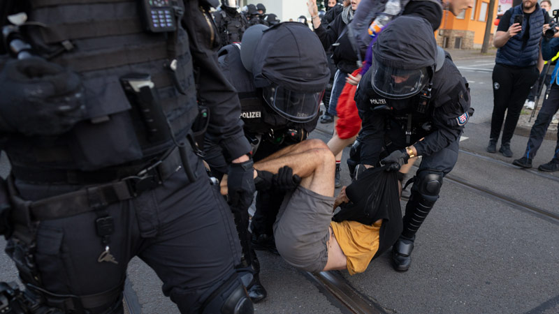Bereitschaftspolizisten in Vollmontur fixieren eine am Boden liegende Person während einer Demonstration; im Hintergrund weitere Beamte und Umstehende, teils mit Kameras.