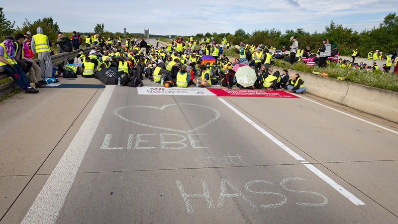 Sitzblockade auf einer Autobahn: Dutzende Personen in gelben Warnwesten sitzen auf der Fahrbahn, auf dem Asphalt ist mit Kreide „Liebe statt Hass“ mit einem Herz geschrieben.