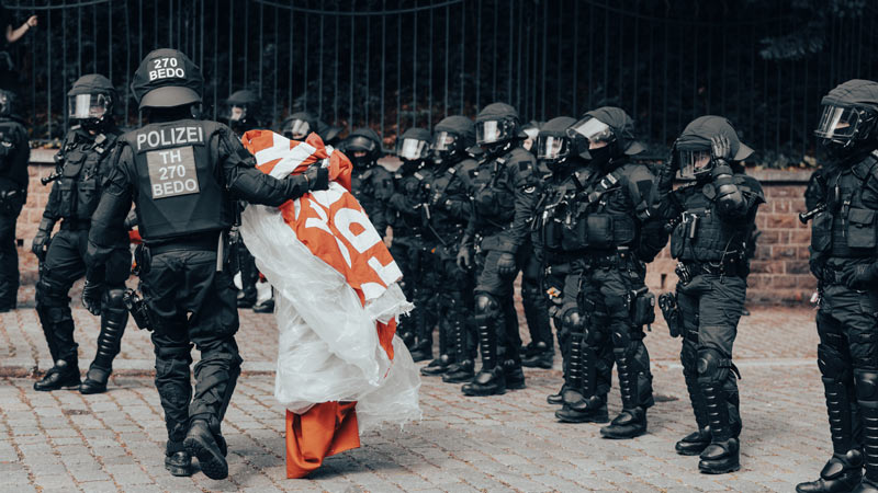 Polizeibeamte in Einsatzausrüstung tragen ein großes orange-weißes Transparent oder Fahnentuch von einem Einsatzort weg, flankiert von einer Reihe weiterer Polizisten.