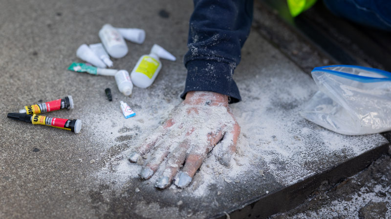Nahaufnahme einer mit weißem Pulver bedeckten Hand auf einem Bordstein, daneben Farbtuben und ein Klarsichtbeutel mit weißem Pulver – Vorbereitung für eine Straßenmarkierung oder Farbaktion.