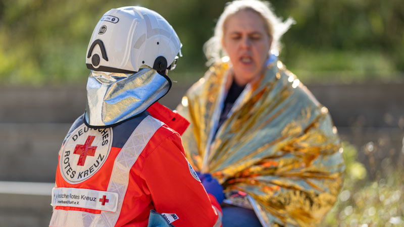 DRK-Rettungshelfer mit Helm im Profil, im Hintergrund eine Frau eingehüllt in eine goldene Rettungsdecke
