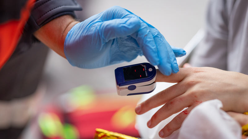 Rettungssanitäter misst mit einem Pulsoxymeter die Sauerstoffsättigung am Finger eines Patienten, Detailaufnahme der Hände