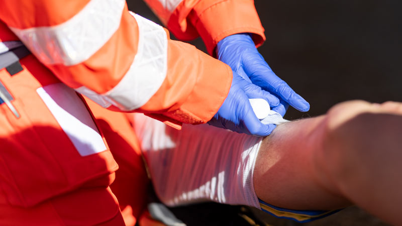 Rettungskräfte des Deutschen Roten Kreuzes legen einem Patienten am Unterarm einen Verband an, Nahaufnahme der Hände in blauen Einmalhandschuhen