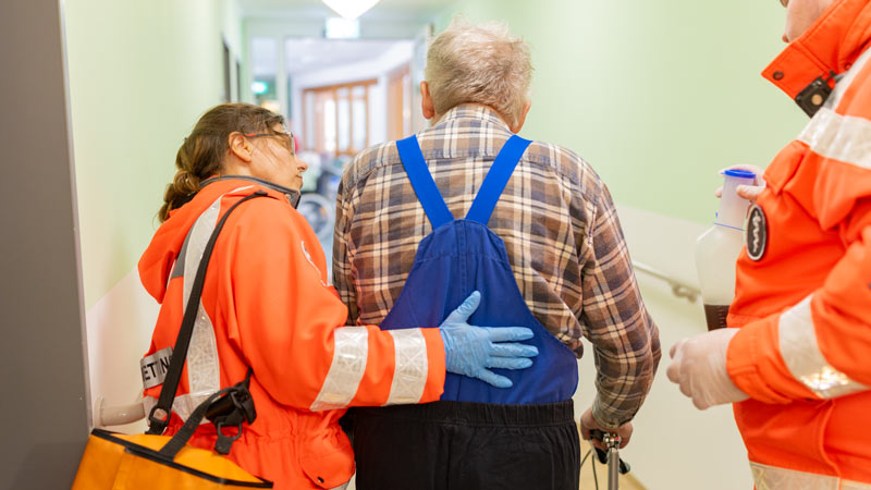 DRK-Helferin stützt einen älteren Bewohner beim Gehen im Flur eines Pflegeheims, im Hintergrund ein weiterer Rettungssanitäter