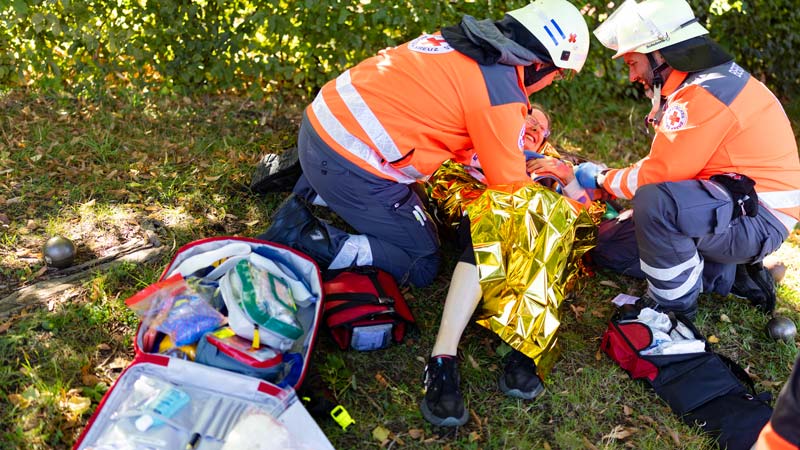 Zwei DRK-Rettungshelfer versorgen einen Patienten in Rettungsdecke auf einer Wiese, im Vordergrund geöffnete Notfallrucksäcke, im Hintergrund Boule-Kugeln