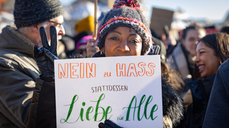 Eine lächelnde Demonstrantin hält ein Schild mit der Aufschrift "NEIN zu HASS, stattdessen Liebe für Alle" während einer Kundgebung.