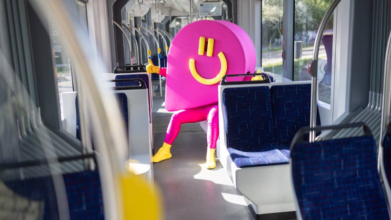 Maskottchen in pinkem REWE Bonus-Kostüm mit gelben Handschuhen sitzt allein in einer modernen Straßenbahn.