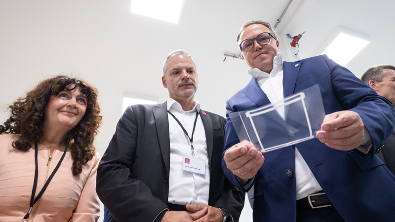 Drei Personen in Businesskleidung betrachten ein transparentes technisches Bauteil in einem hellen Raum.
