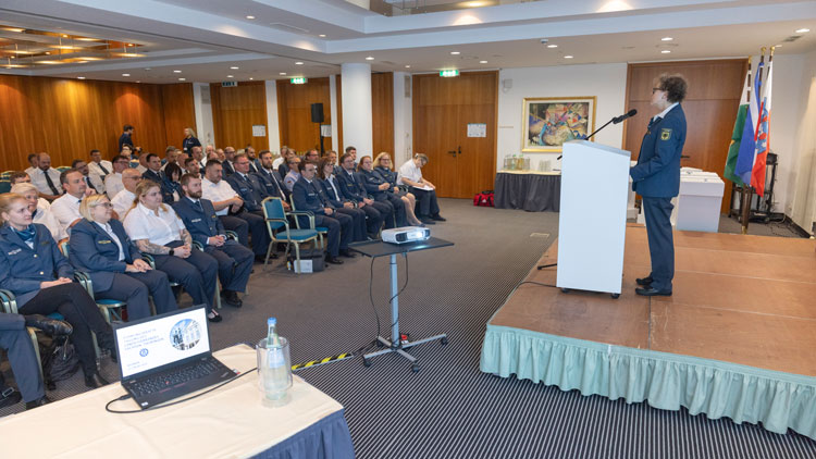 Eine Frau in Uniform steht auf einer Bühne und hält eine Präsentation vor einem Publikum. Das Publikum besteht überwiegend aus Personen in Uniform, die aufmerksam zuhören. Im Raum befinden sich eine Projektionsleinwand und eine Laptop-Präsentation im Vordergrund. Im Hintergrund hängen mehrere Flaggen, und rechts ist ein kleiner Tisch mit Getränken zu sehen.