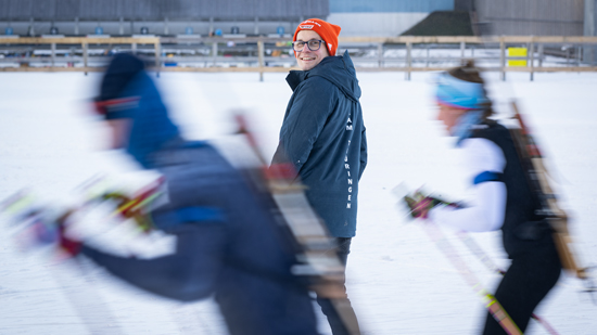 Eine lächelnde Person mit Brille und orangefarbener Mütze steht im Vordergrund, während im Hintergrund verschwommene Skiläufer vorbeiziehen.